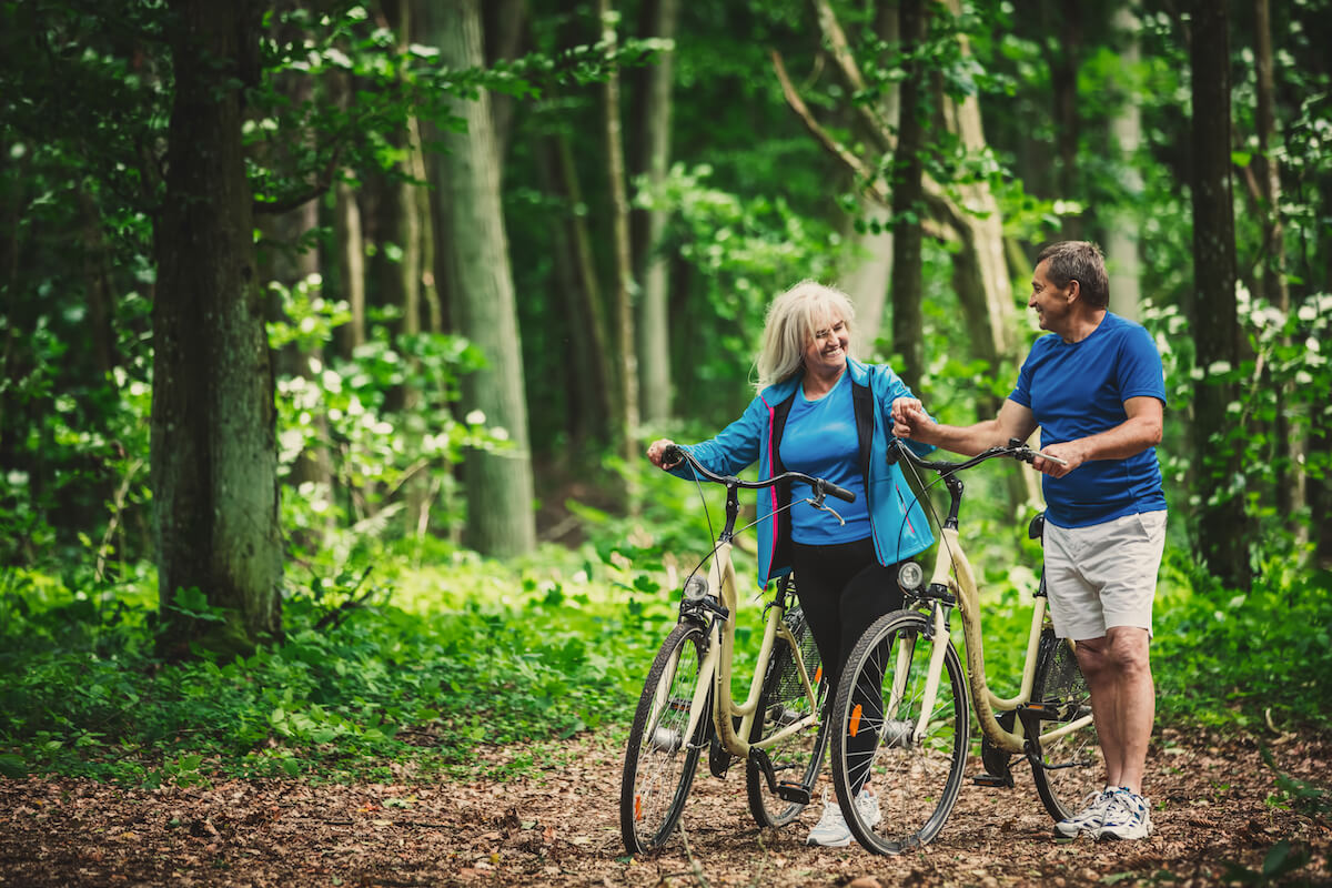 Retired Couple Walking With Bikes In The Forest.